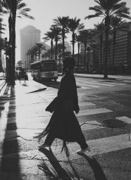 Black and white editorial photo of model Caren Jepkemei walking across a sunlit city crosswalk lined with palm trees.