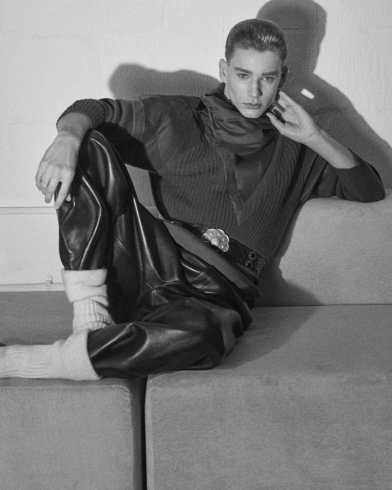 Monochromatic portrait of a stylish adult male wearing leather pants, a V-neck sweater, and a scarf, striking a pose in a studio setting.