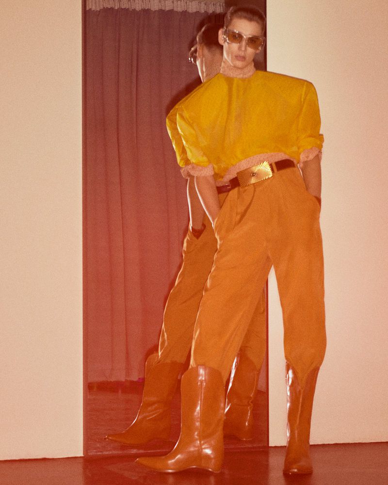 Person in yellow crop top, orange pants, and cowboy boots poses near a mirror. Fashion shot with retro vibe and indoor setting.