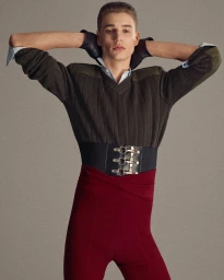 Model Cas Lambrechts posing with arms behind head wearing a ribbed sweater, wide buckled corset belt, and deep red pants.