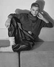 Model Cas Lambrechts seated on a modular sofa wearing a ribbed sweater and leather trousers in a black and white fashion editorial.