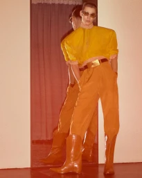 Model Cas Lambrechts wearing a yellow padded top, orange trousers, and tan leather boots posing in front of a full-length mirror.