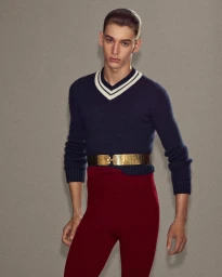 Model wearing a navy V-neck sweater, a gold metallic belt, and red trousers against a grey textured background.