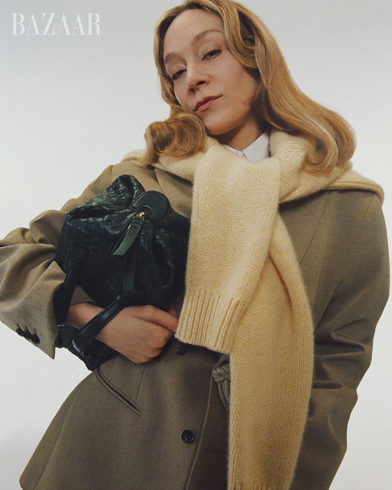 Blonde woman in beige coat and cream scarf holds black handbag in a studio portrait for Bazaar magazine, fashion photography.