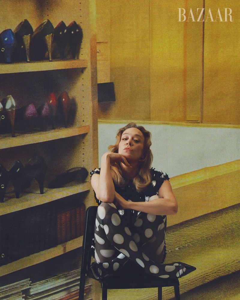Young woman poses while sitting in a chair in front of a shelf. The woman wears polka dot tights and a dark top. Bazaar logo visible.