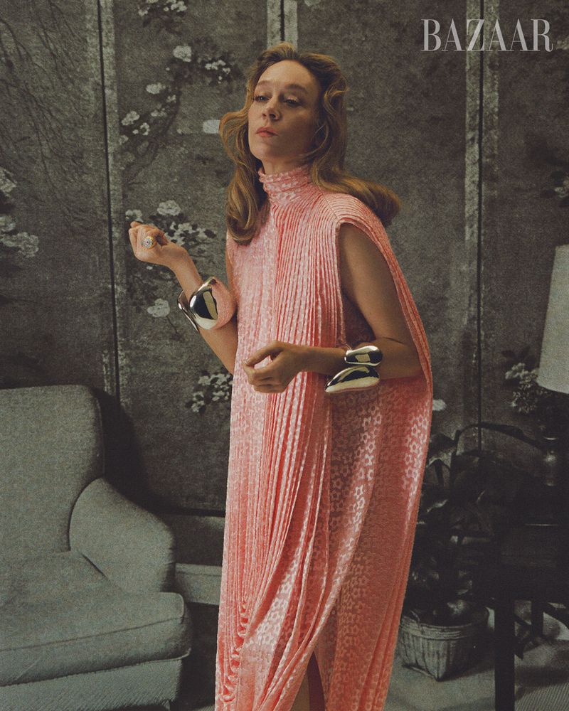 Woman in a flowing peach gown, wearing large silver cuff bracelets, poses in front of a floral wallpaper, captured in a fashion editorial.
