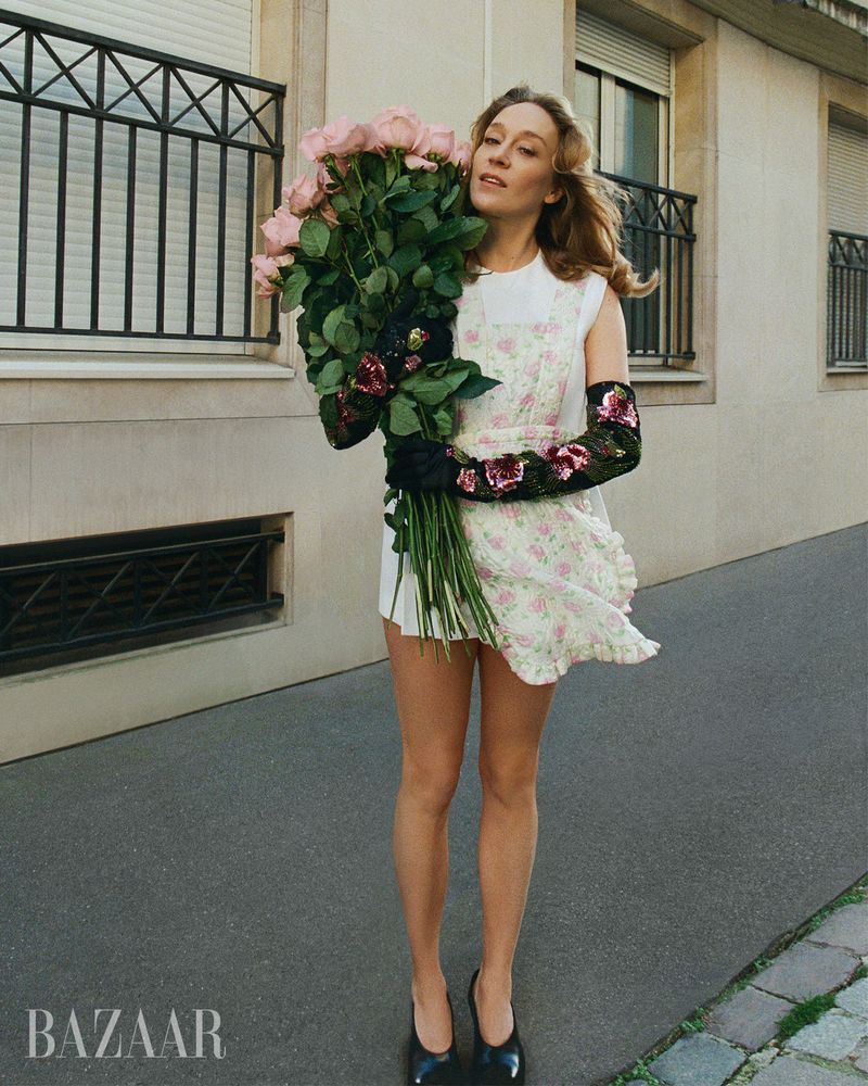 Adult female fashion model in short white floral dress poses with large bouquet of pink roses on city sidewalk near building.