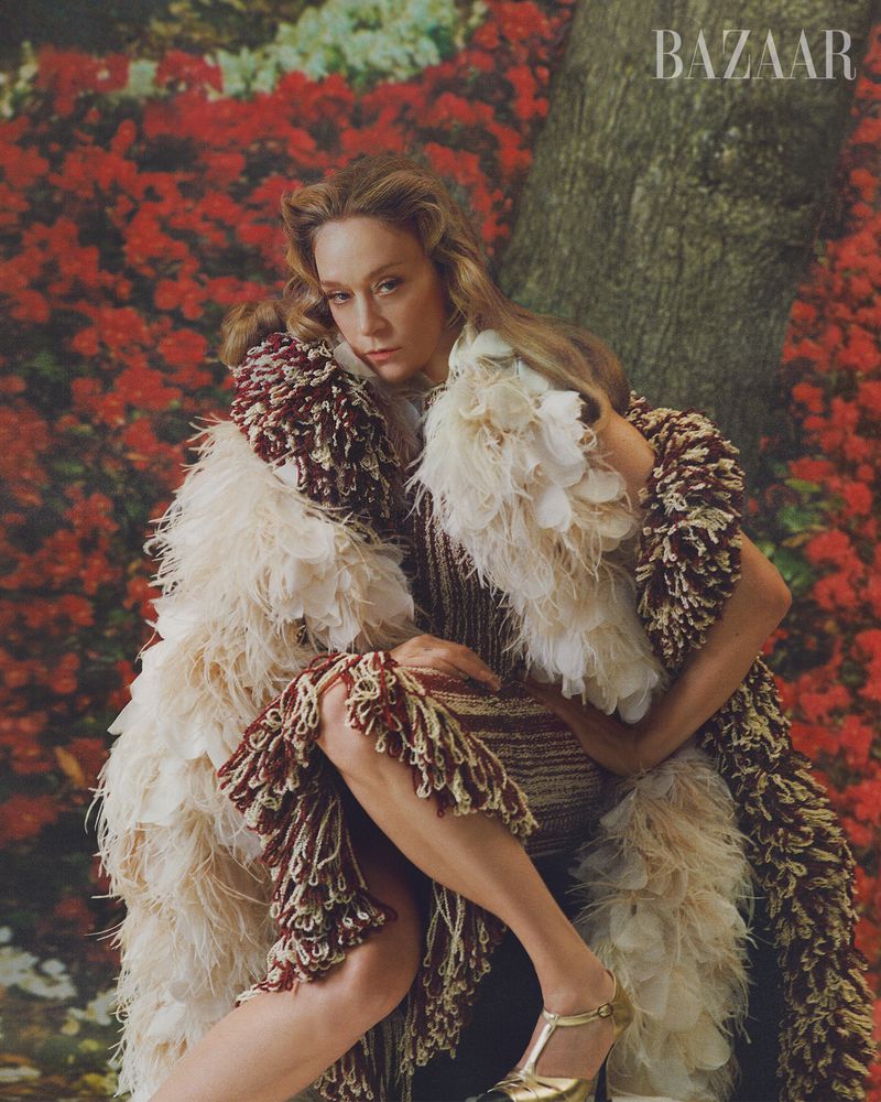 A woman poses in a feathered and fringed dress against a backdrop of flowers and a tree trunk, with the 'BAZAAR' logo visible.