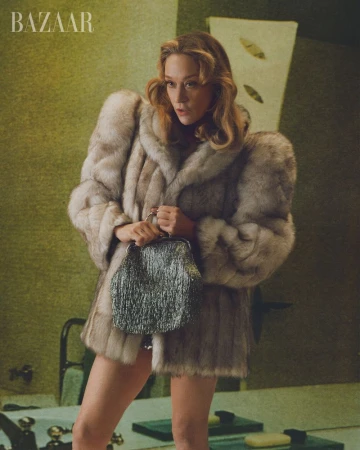 Chloë Sevigny posing in an oversized plush fur coat while holding a textured metallic clutch bag in a green interior.