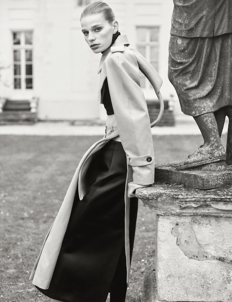 Monochrome fashion shot of an adult woman in a trench coat leaning against a weathered stone structure in front of a building.