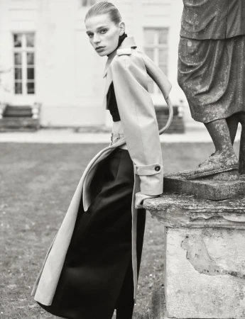 Monochrome fashion shot of an adult woman in a trench coat leaning against a weathered stone structure in front of a building.