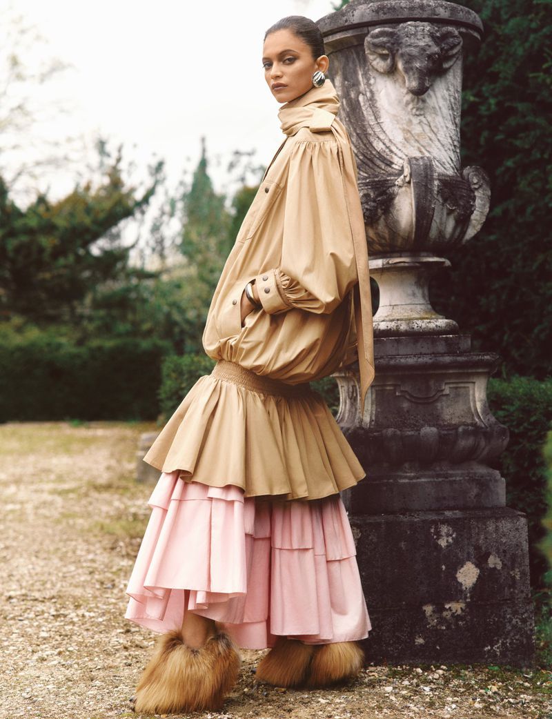 A person wearing a large beige jacket and pink ruffled skirt poses next to a large stone vase in an outdoor setting with green trees.