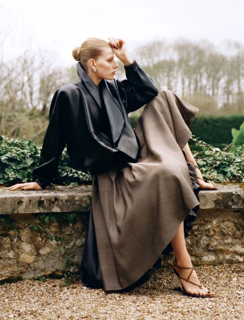 Fashion shot of woman in black jacket and brown skirt sitting on stone wall in outdoor setting with greenery in the background.