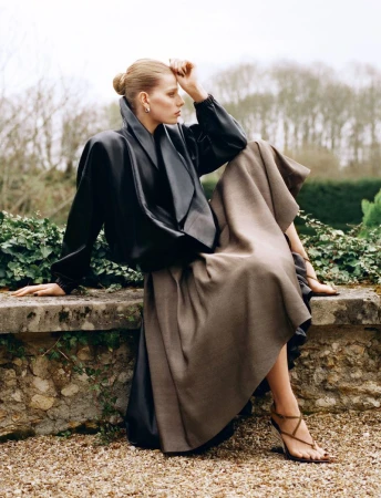 Fashion shot of woman in black jacket and brown skirt sitting on stone wall in outdoor setting with greenery in the background.