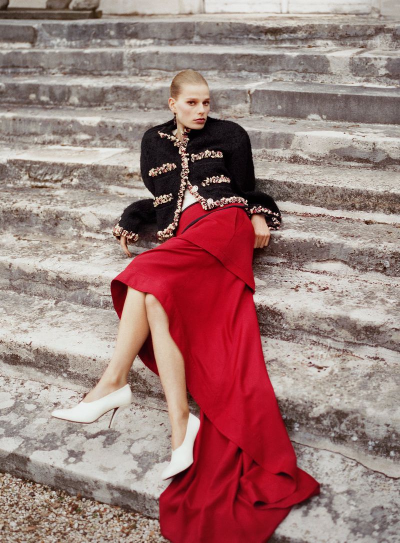 Fashion shot of an adult female model wearing a black jacket and a red skirt sitting on concrete steps in an outdoor environment.