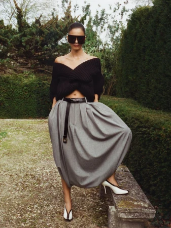 Woman in an off-the-shoulder black sweater, gray balloon skirt, and slingback heels poses in an outdoor garden setting with green foliage.
