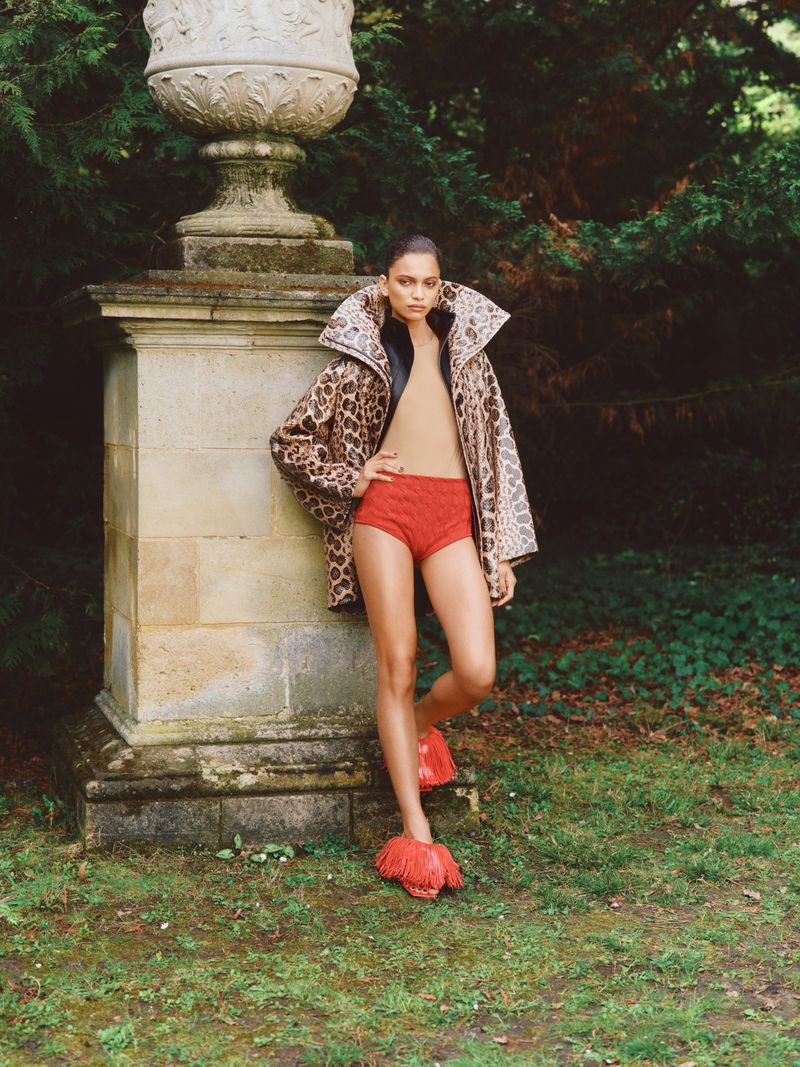 A stylish adult female poses outdoors wearing a leopard-print coat, red shorts and shoes, next to a weathered stone pedestal. 