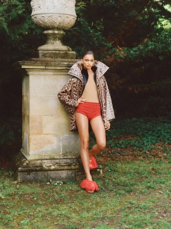 A stylish adult female poses outdoors wearing a leopard-print coat, red shorts and shoes, next to a weathered stone pedestal. 