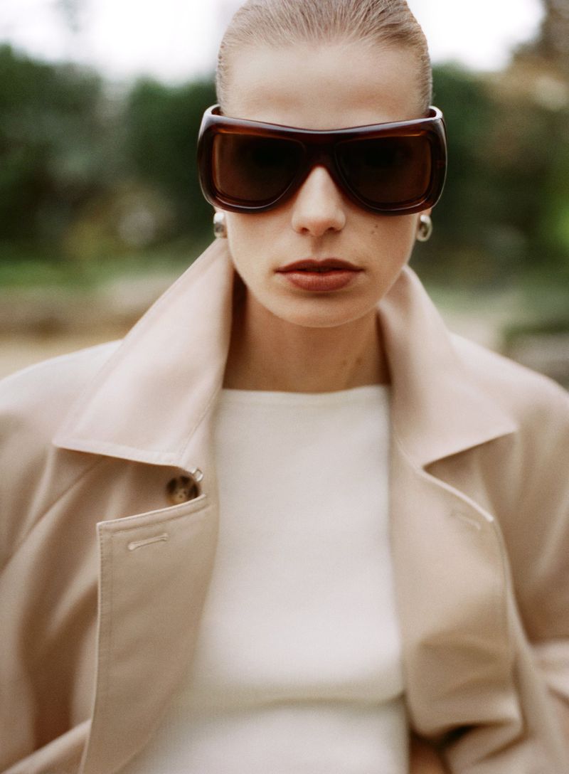 Close-up of a woman in a tan coat and brown sunglasses with a blurred outdoor backdrop. Beauty, fashion, and style are emphasized.