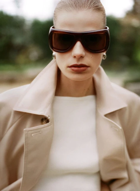 Close-up of a woman in a tan coat and brown sunglasses with a blurred outdoor backdrop. Beauty, fashion, and style are emphasized.