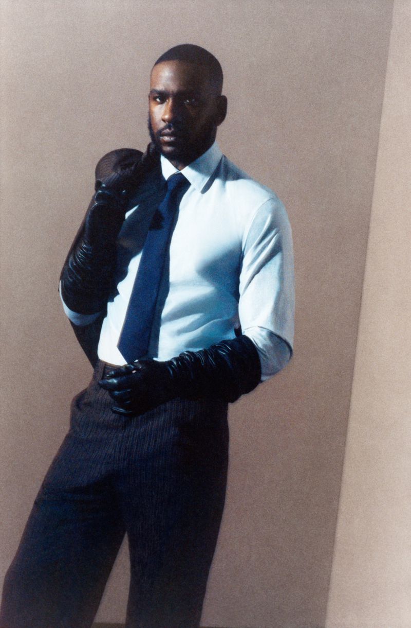 Portrait of a stylish Black man in dress shirt, dark tie, and gloves poses against a neutral backdrop with a jacket over his shoulder.