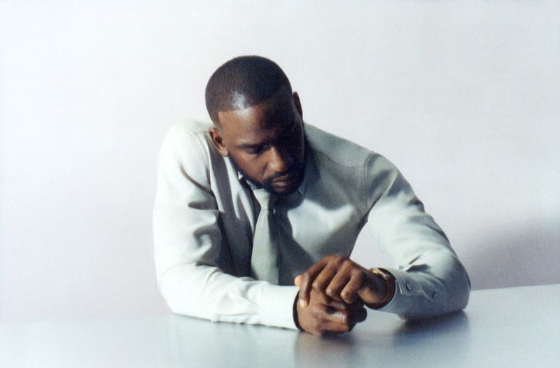 Adult Black man sits at a table with his hands clasped, wearing a pale-grey shirt and tie in an indoor setting against a white backdrop.