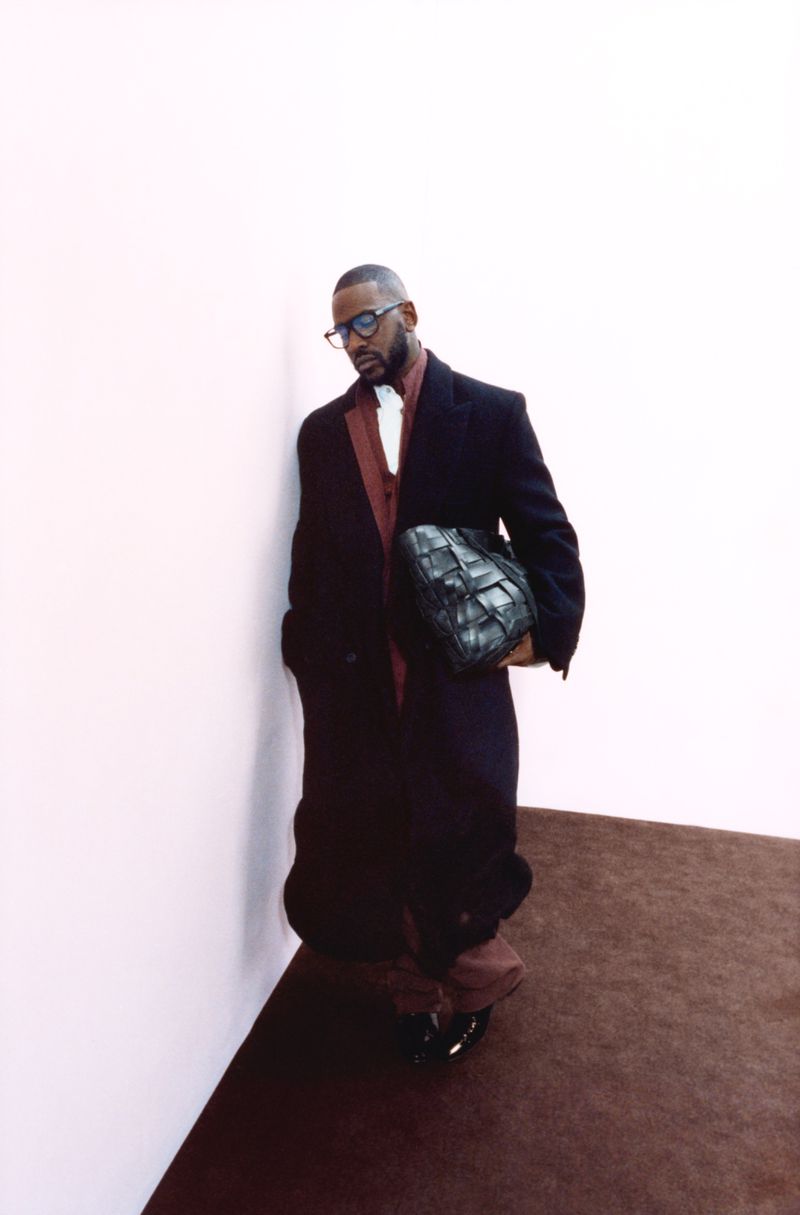 Adult male in a black overcoat and glasses indoors, holding a woven black bag, with neutral white wall and brown carpet background.
