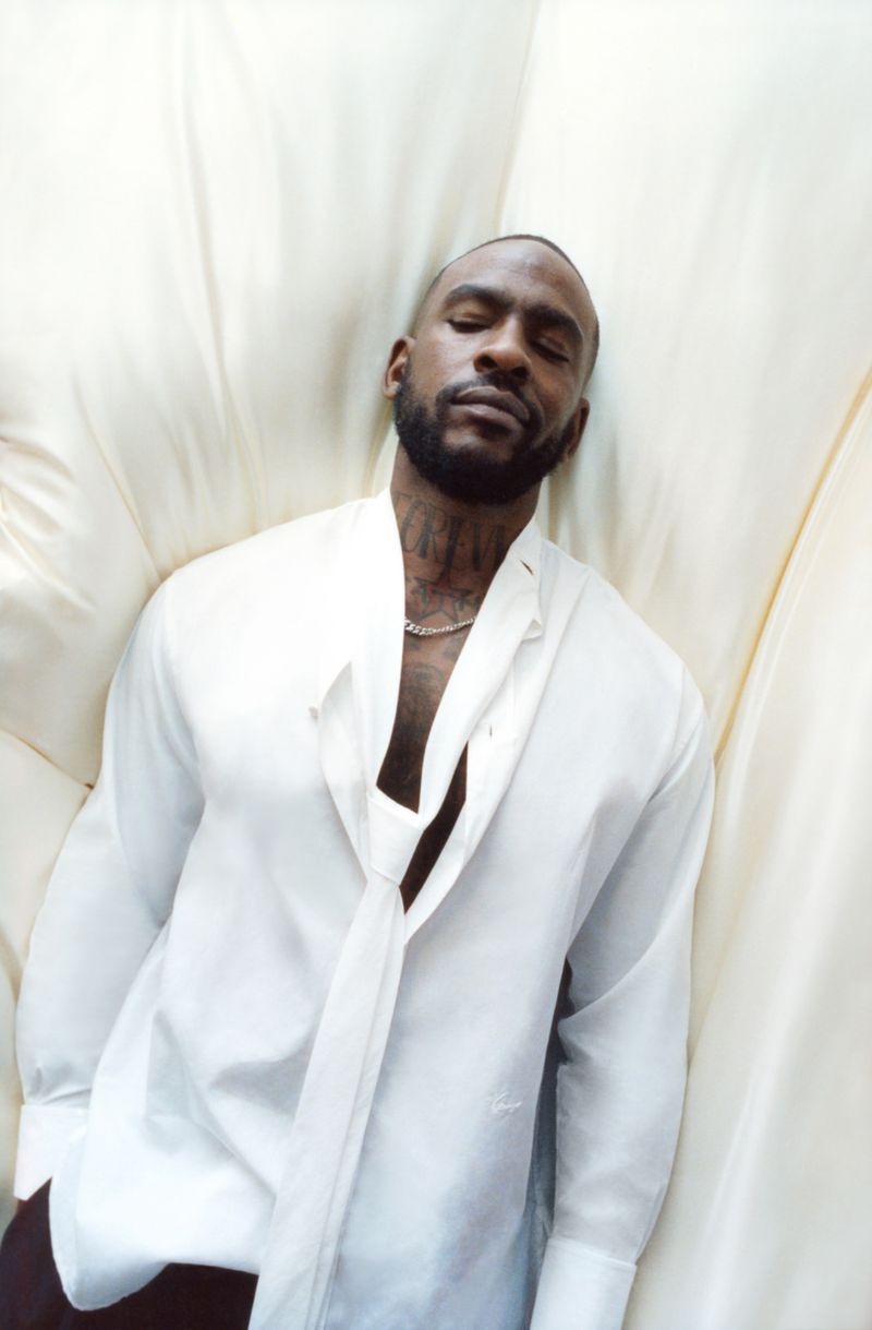 A man with dark hair and a beard is lying down in a white collared shirt with an unbuttoned collar, against an off-white backdrop.