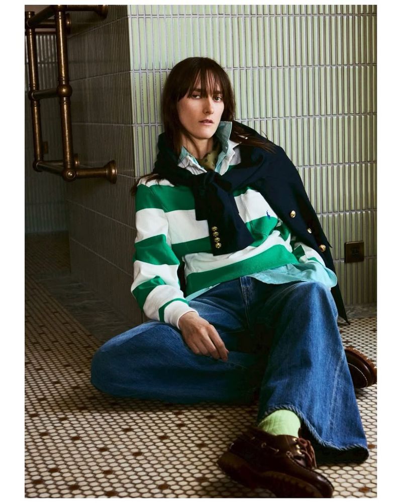 Stylish woman poses indoors wearing a striped sweater, jeans, and leather boots. Tiled walls and floor create a casual, fashionable scene.