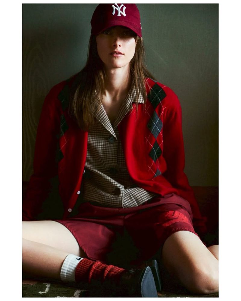 Model Sveta Kare wearing a red argyle cardigan, collared shirt, and baseball cap seated on the floor for Elle Japan.