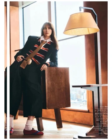 Woman in black suit, striped shirt, and red shoes poses indoors with a vintage lamp and wooden table against a window backdrop.
