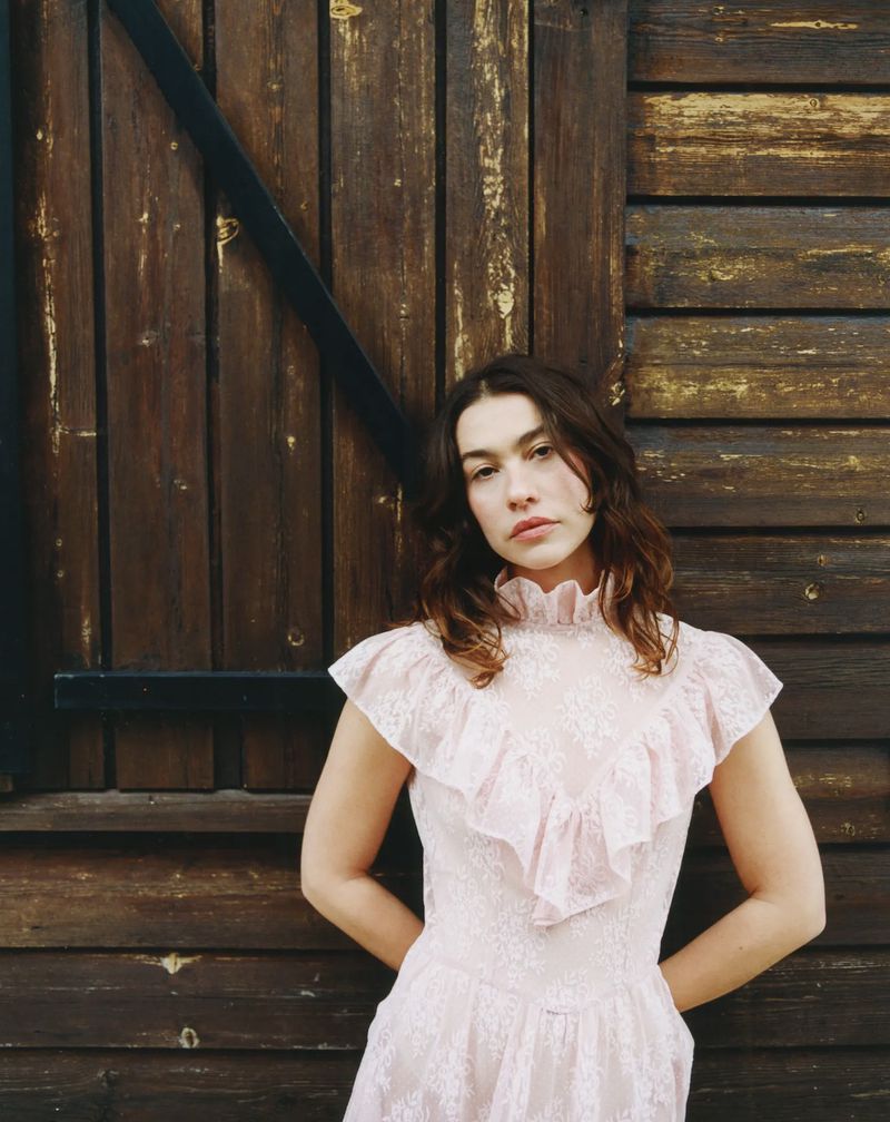 Young woman with brunette hair posing in a pink lace dress in front of an old wooden door in a medium shot outdoors.