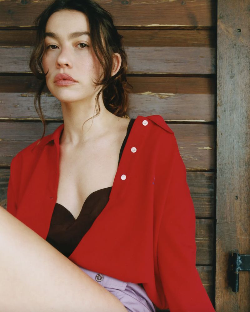 Portrait of a woman with dark hair wearing a red shirt and lavender shorts, posing against a weathered wooden background.