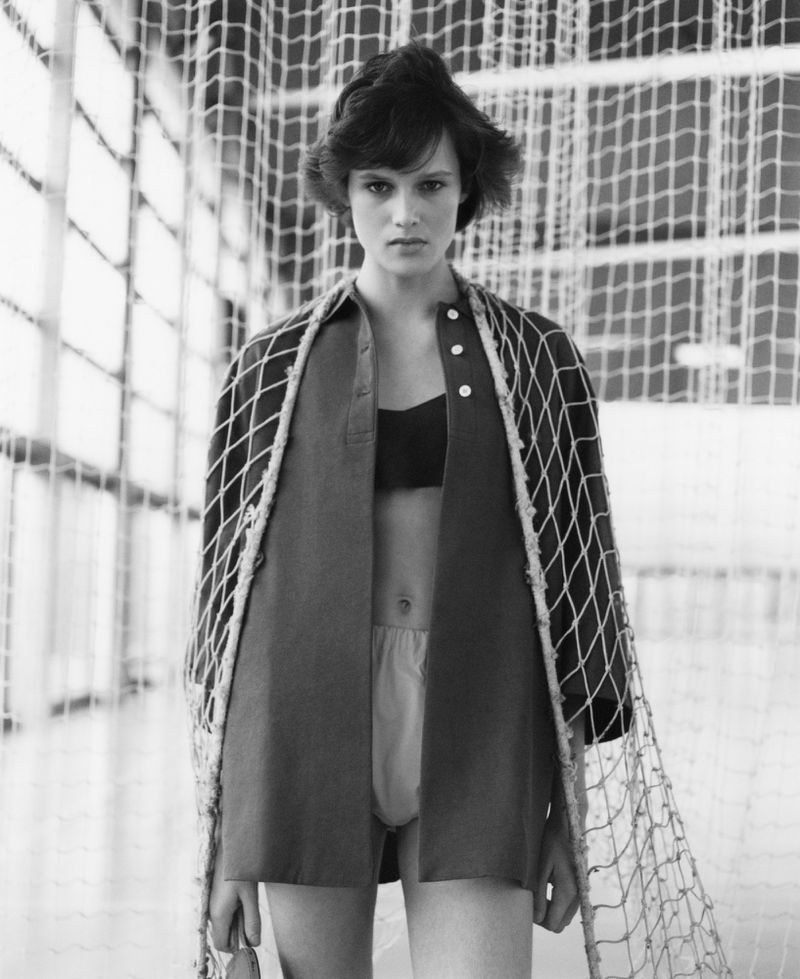 Black and white portrait of a woman with short hair wearing a bra and an open shirt draped with netting in front of a net background.