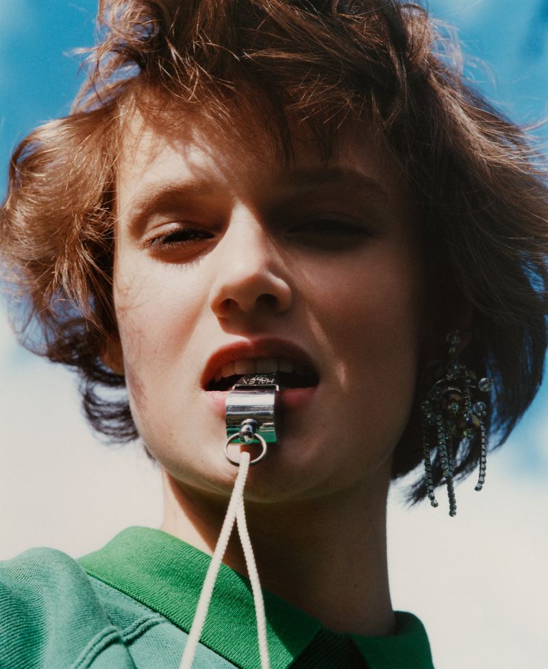 Close-up of a woman with short brown hair wearing a green shirt, earrings and holding a metal whistle in her mouth, against a blue sky.