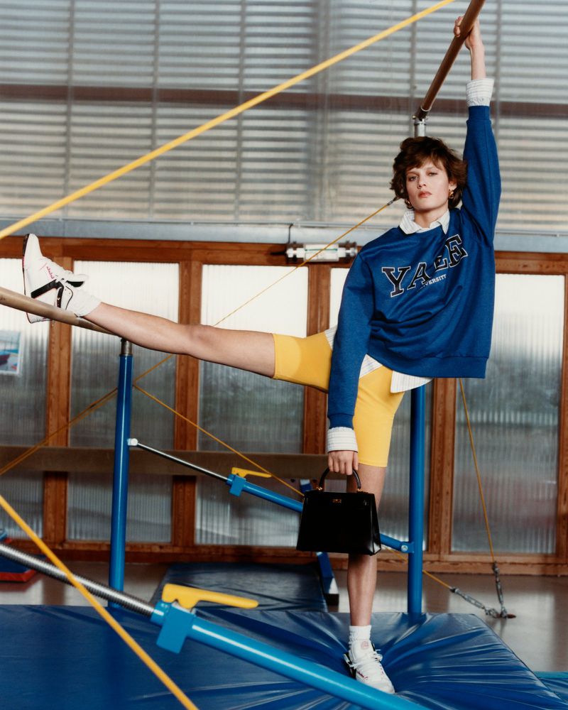 A woman in a Yale sweatshirt stretches on gymnastic bars in a gym setting, holding a black handbag, showcasing a fashion editorial style.