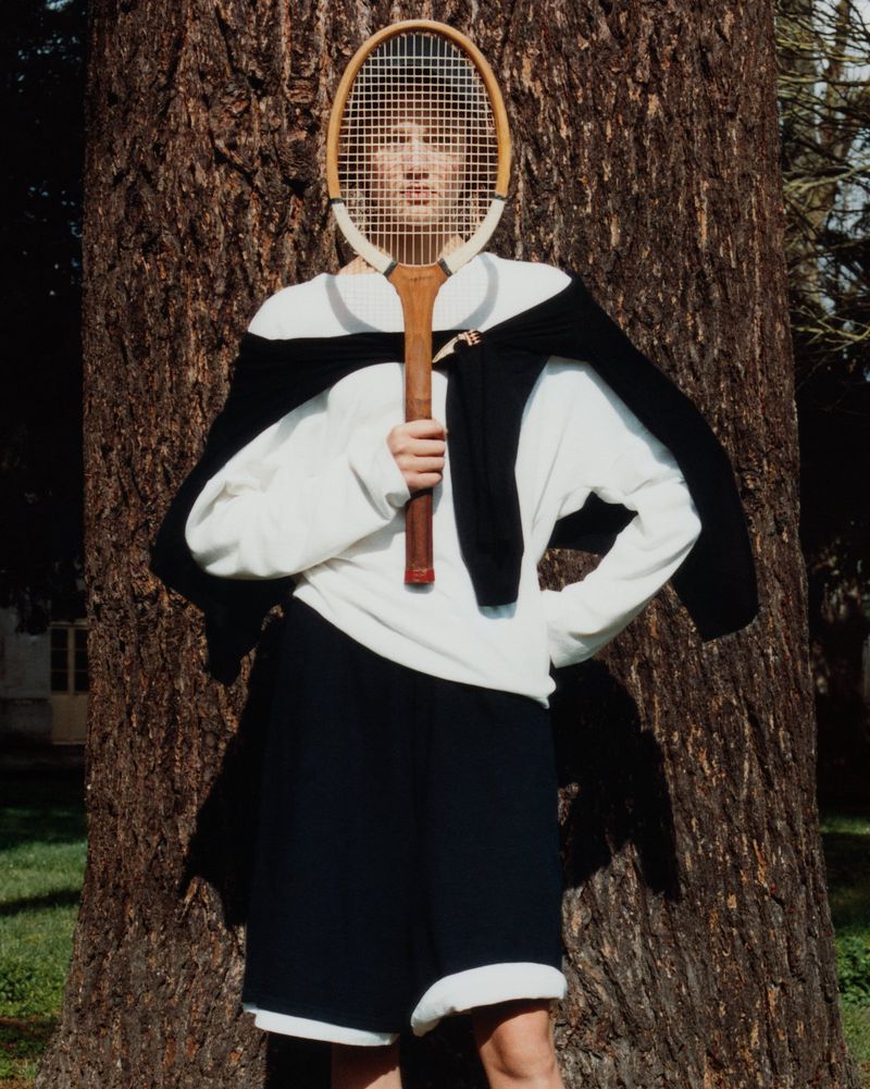 Person stands in front of tree holding a vintage tennis racket over their face, wearing a white sweatshirt and black shorts outdoors.
