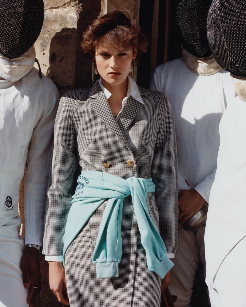 Woman in checkered blazer with blue sweater around her waist, posed next to a group of fencers in white outfits and black masks.