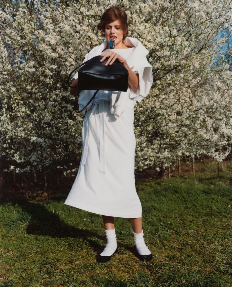 An adult female model poses in a white dress and holds a black bag in a sunny outdoor setting with flowering trees in the background.