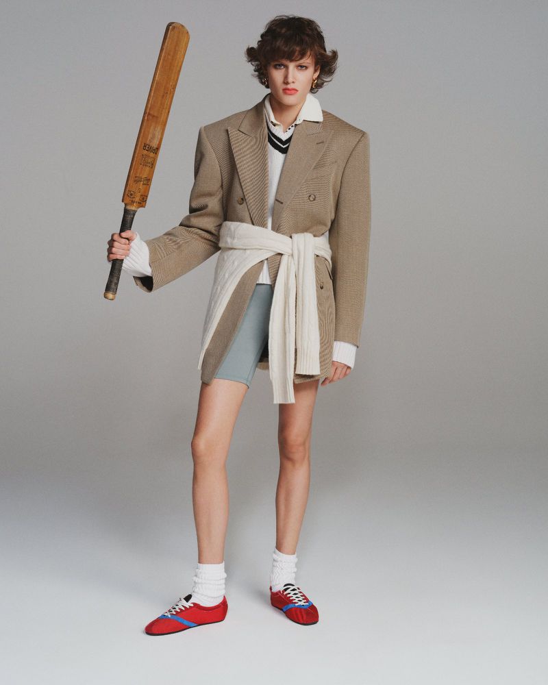 Woman in a blazer and athletic shorts holding a cricket bat in a fashion studio shot with athletic wear and neutral colors.
