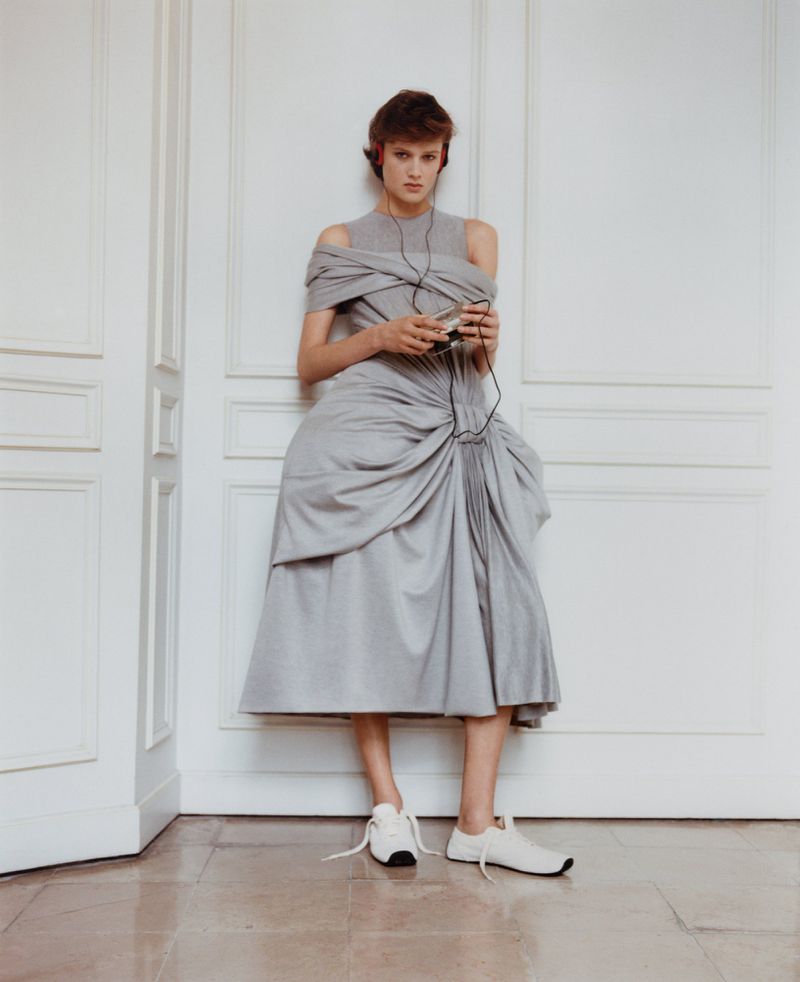 A woman poses indoors wearing a grey dress with a modern design, white sneakers, and headphones while holding an electronic device.