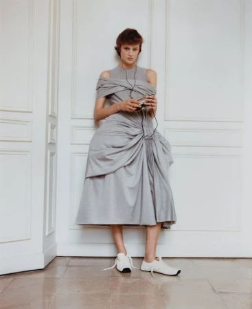 A woman poses indoors wearing a grey dress with a modern design, white sneakers, and headphones while holding an electronic device.