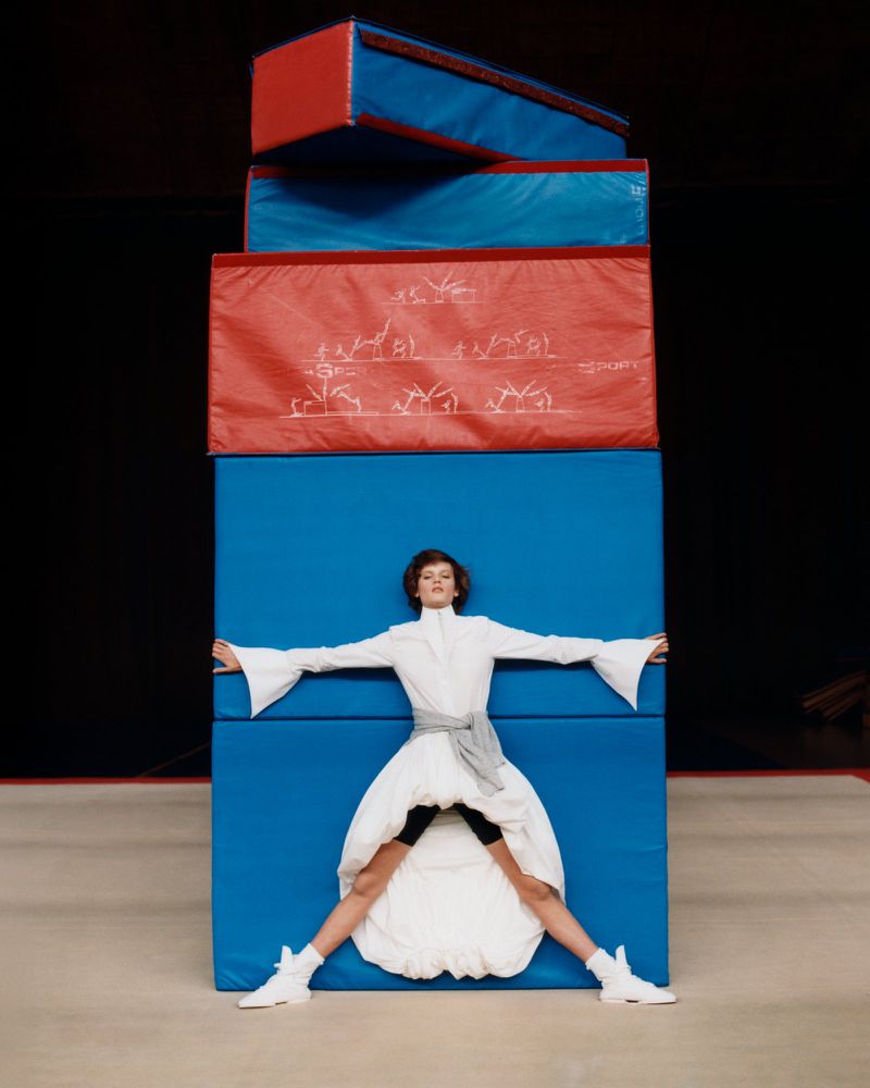 A woman in a white dress poses dramatically in front of stacked blue and red mats in a minimalist photo studio setting with dark background.