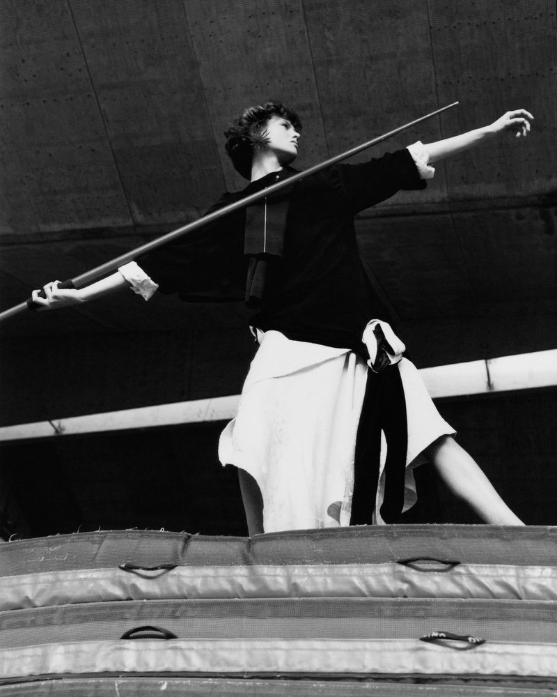 Monochrome shot of a person in fashionable attire, holding a long pole, standing atop stacked mats against a grey concrete backdrop.