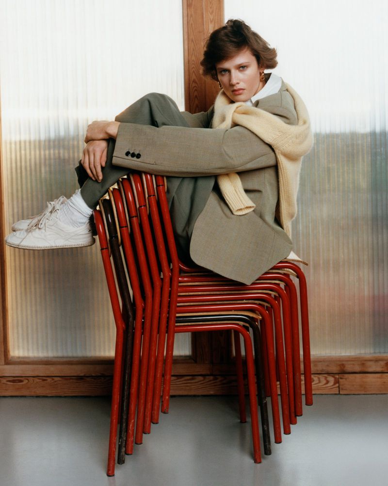 A woman with short brunette hair sits atop a stack of red metal chairs wearing a tan blazer, green trousers, a cream-colored sweater and white sneakers.