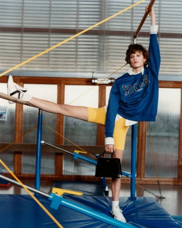 Model in blue sweatshirt and yellow shorts posing on gymnasium equipment holding a black handbag