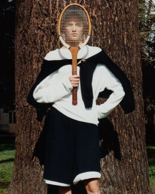 Model holding a vintage wooden tennis racket in front of their face, wearing a white shirt and black sweater against a tree trunk.