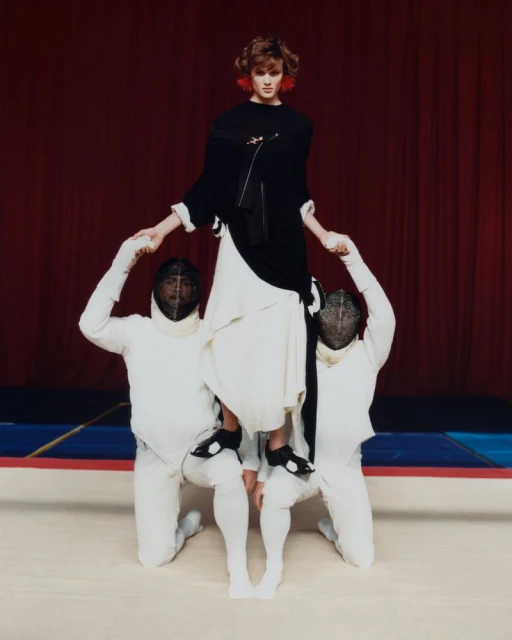 Model Noor Khan stands supported by two fencers in white fencing uniforms and mesh masks against a deep red curtain.