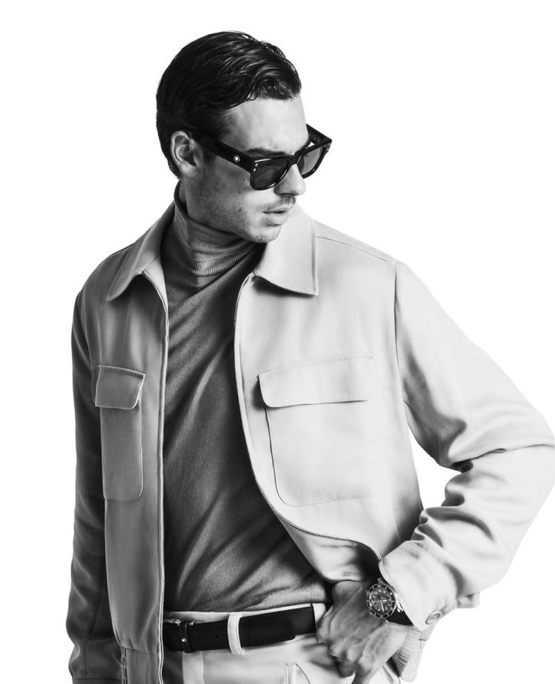 Monochrome portrait of a man in sunglasses and a tan jacket looking away against a white background in a studio shot.