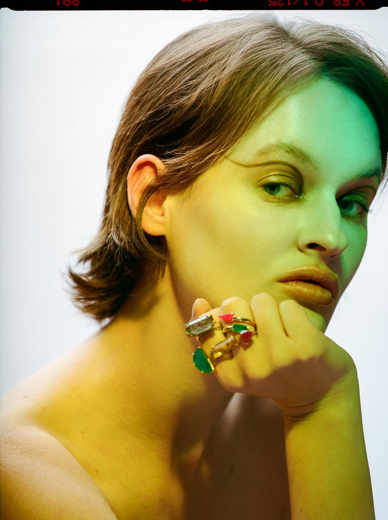 Close-up of a young fair-skinned woman with short brown hair, wearing gold rings with green, red, and white gems, against a plain white background.